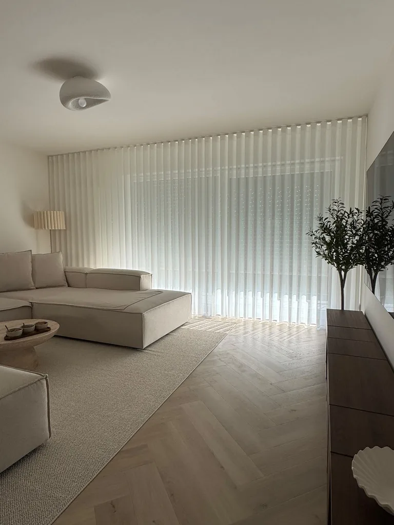 Weiße transparente Gardinen im hellen Wohnzimmer mit beigem Ecksofa und Fischgrätboden