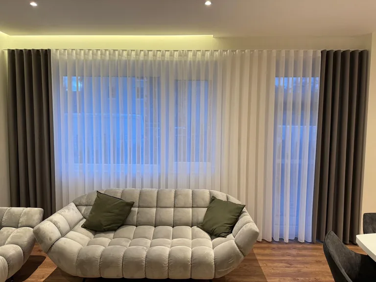 Weiße Gardinen und dunkle Seitenschals hinter einem hellen Sofa mit grünen Kissen
