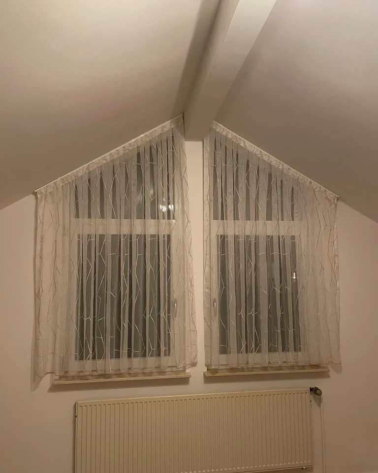 Weiße gemusterte Gardinen an einem Giebelfenster über dem Heizkörper, Frontalansicht