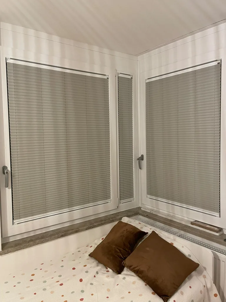 Geschlossene helle Plissees an einer Eckfensterfront über einem Bett
